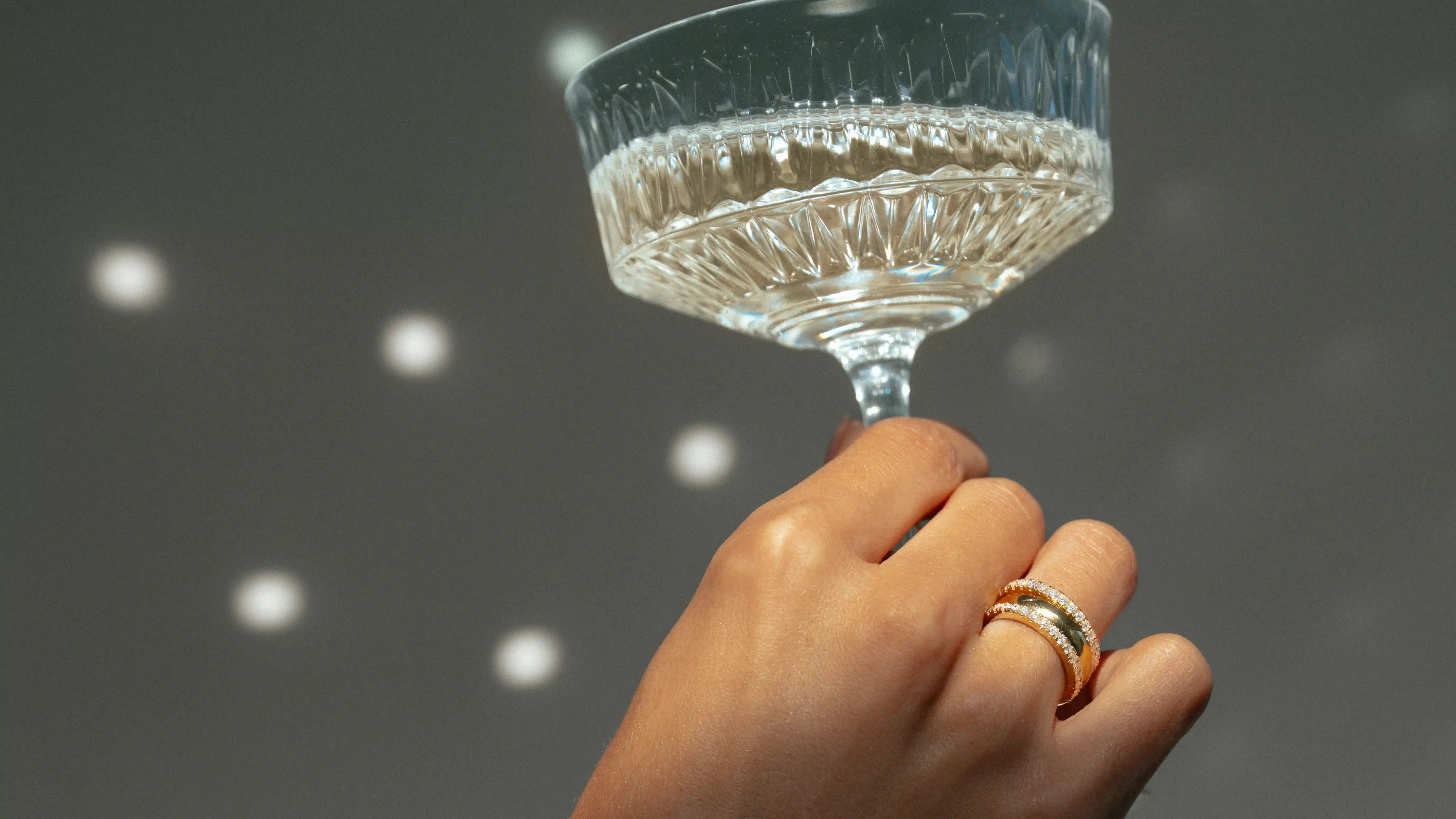 Hand met 18 karaat gouden ringen met lab grown diamanten houdt een glas champagne vast.